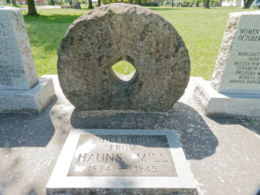photo shows a large mill stone between two memorial stones.  In front of it is a stone that reads " Millstone from Hauns Mill"